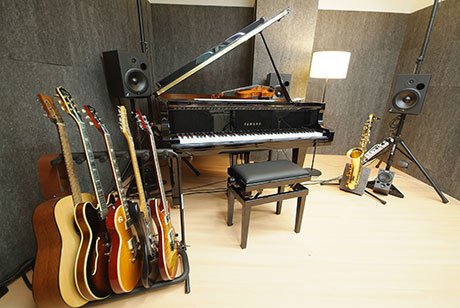 Music studio with a grand piano in the center, a violin placed on top, a guitar stand on the left, two speakers on stands to the right, a saxophone on the floor, and soundproofing panels on the walls.