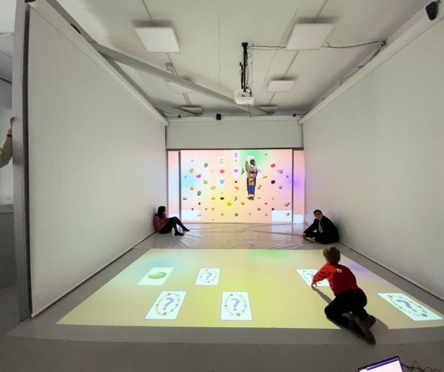 Interactive exhibition space with projections on the floor and wall: a person is suspended in front of a colorful climbing wall, while other people, including a child, interact with the projected images on the floor.