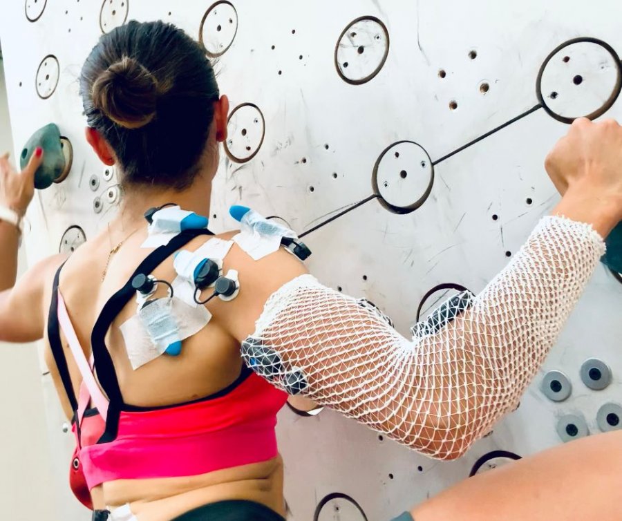 Person seen from behind climbing an interactive indoor wall, with sensors and straps attached to shoulders and back.