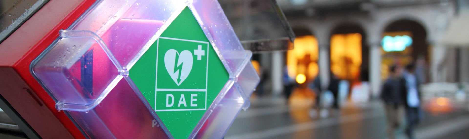 An automated external defibrillator placed outdoors in a transparent case with green signage, located in an urban square with historic buildings in the background and people walking.