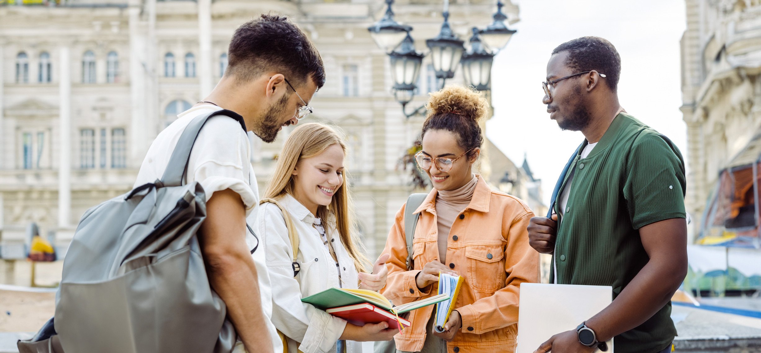 Gruppo di quattro studenti internazionali che parlano e sorridono all’aperto in una piazza europea, con edifici storici sullo sfondo.