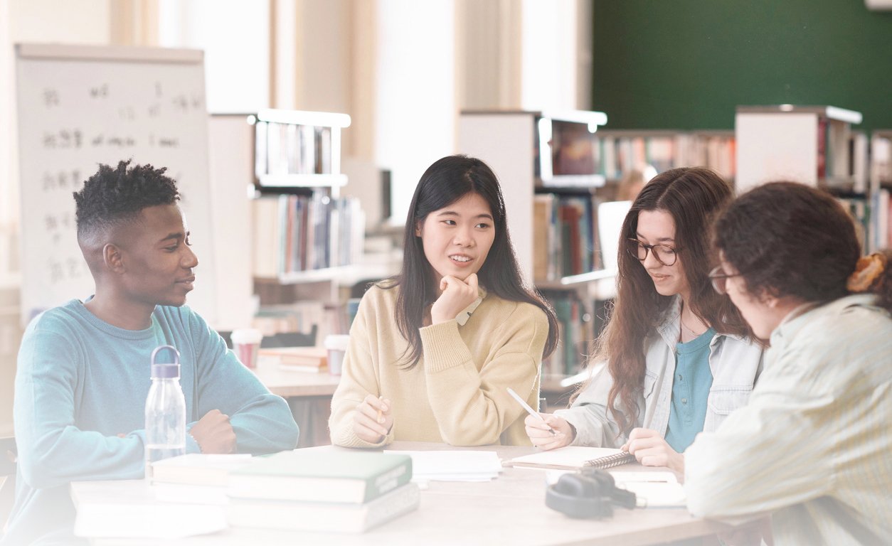 Group of diverse young students discussing and sharing ideas in a library or study environment.